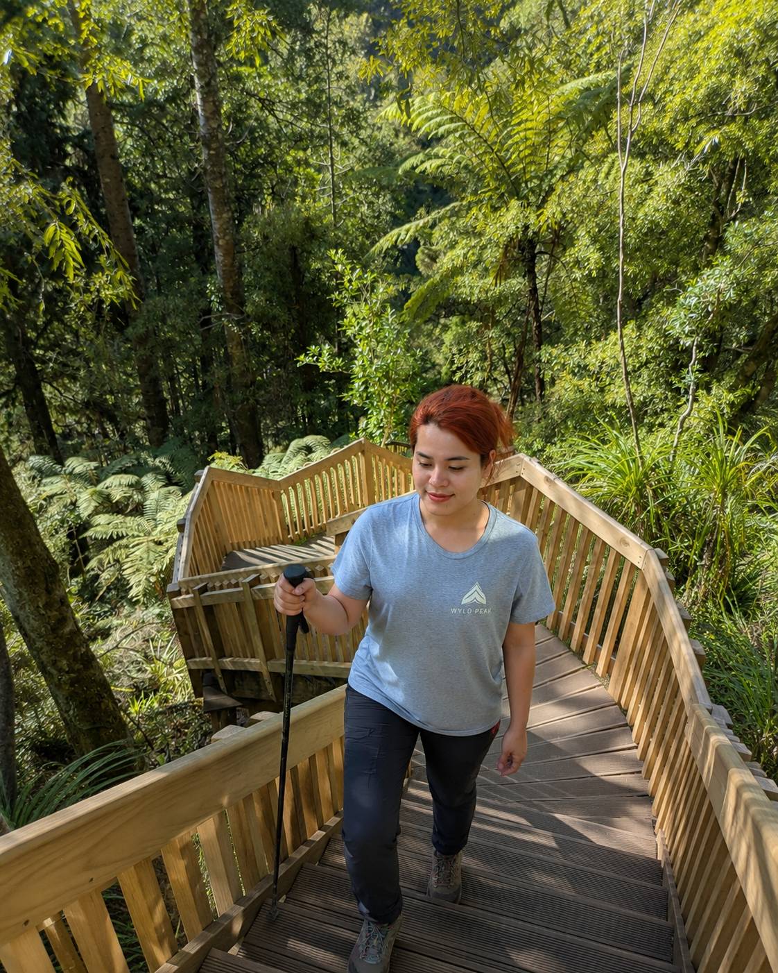 A woman with short red hair wearing a light blue t-shirt and dark pants walks down a wooden boardwalk bridge surrounded by a lush green forest.