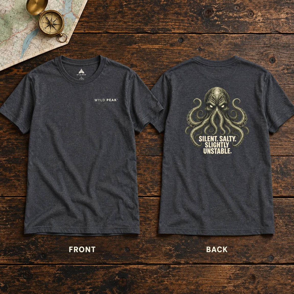 Two dark heather gray t-shirts shown flat on a rustic wooden surface, displaying the front and back designs. The front features a small white 'WYLD PEAK' logo on the left chest, while the back features a large olive-green octopus graphic with the text 'SILENT, SALTY, SLIGHTLY UNSTABLE.