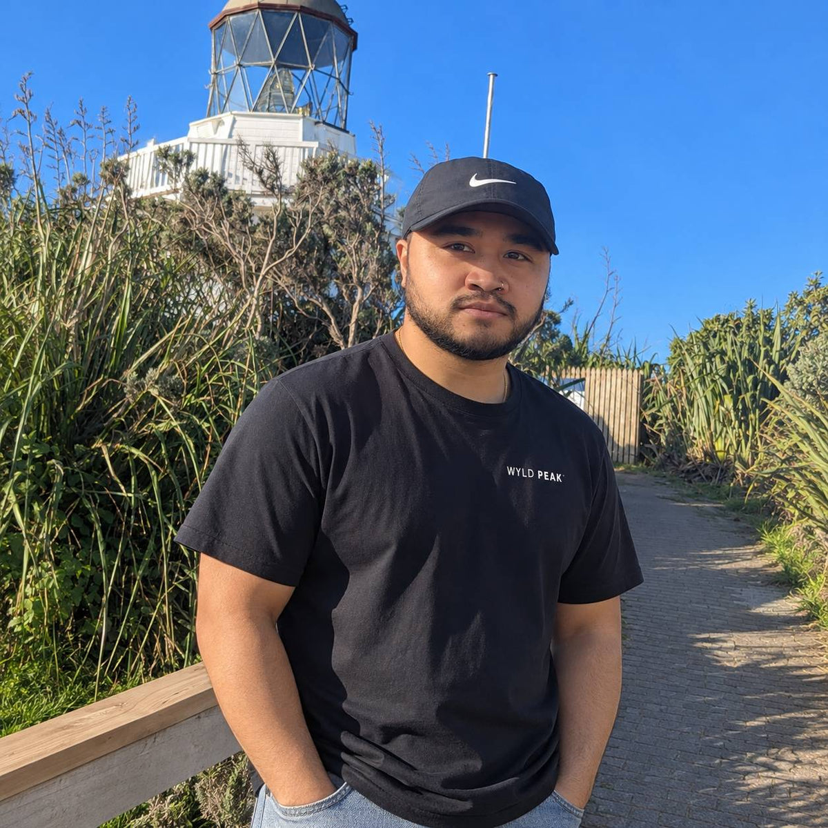 A man wearing a black t-shirt with 'WYLD PEAK' printed on the chest and a black Nike cap, standing outdoors on a path with a lighthouse and greenery in the background.