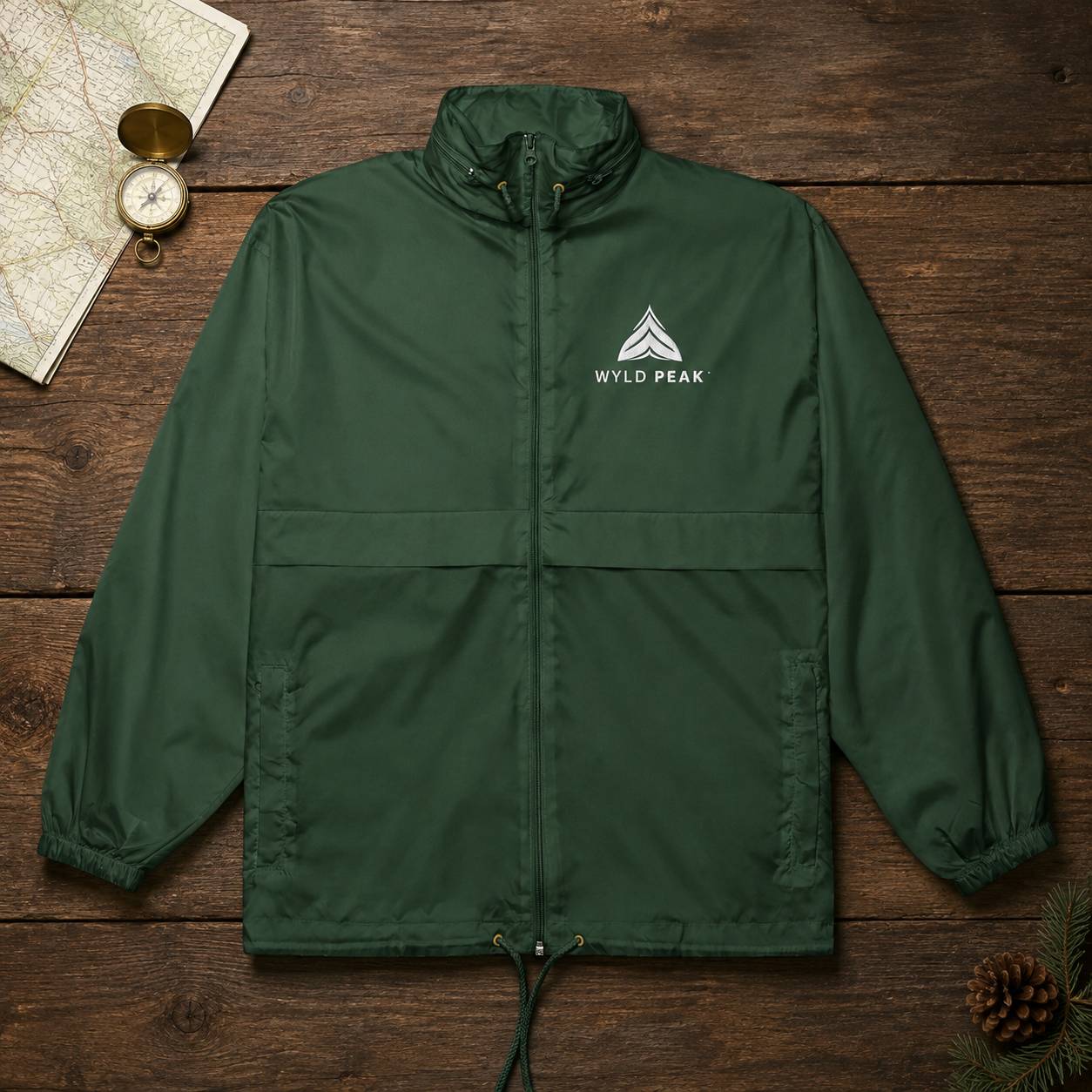 A dark green lightweight jacket with a white 'WILD PEAK' logo on the left chest, laid flat on a rustic dark wood background with a map, compass, and pinecone props.