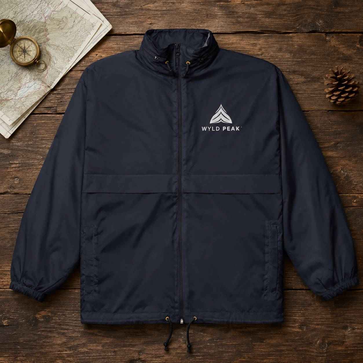 A dark navy blue lightweight jacket with a white 'WYLD PEAK' logo on the left chest, laid flat on a rustic wooden surface with a compass and map in the top-left corner and a pine cone on the right.