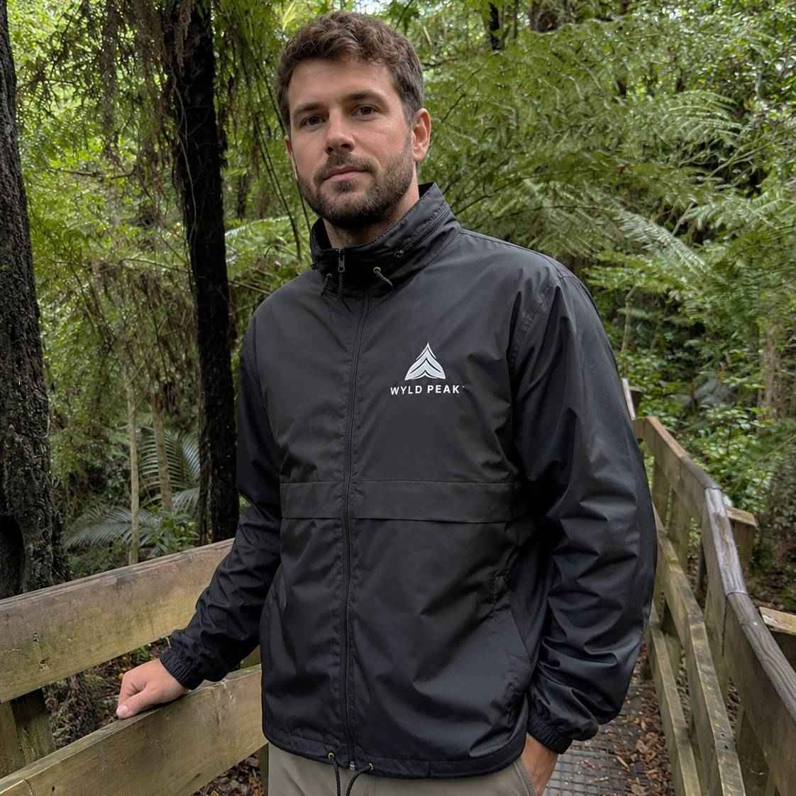 A man wearing a black waterproof jacket with a white 'WILD PEAK' logo on the chest, standing on a wooden boardwalk in a lush green forest.