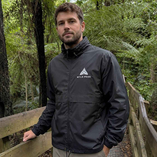 A man wearing a black waterproof jacket with a white 'WILD PEAK' logo on the chest, standing on a wooden boardwalk in a lush green forest.