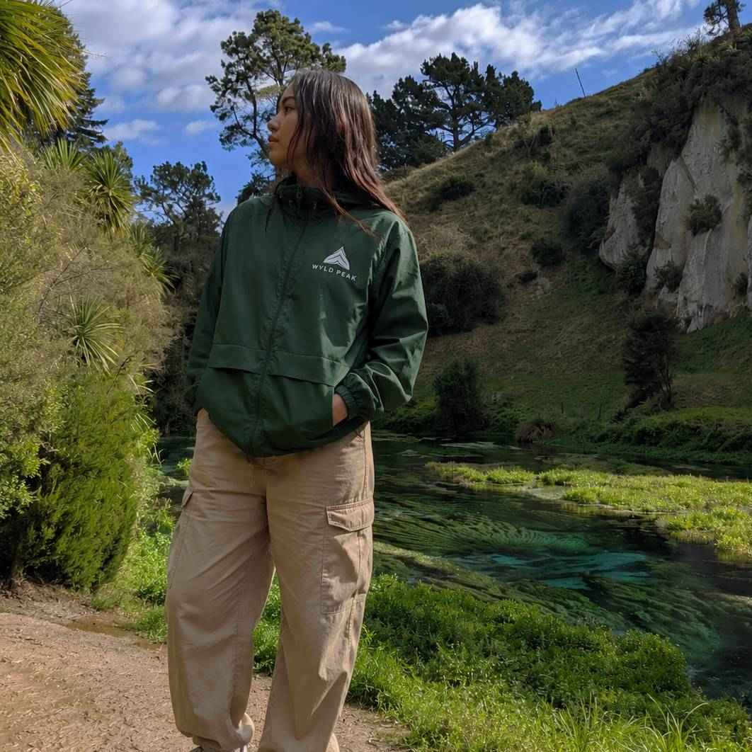 A woman wearing a dark green zip-up hooded jacket with a white 'WILD PEAK' logo and beige cargo pants, standing outdoors in a scenic landscape with a river, green hills, and trees under a blue sky.