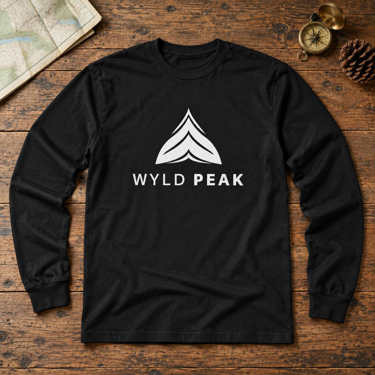 A black long-sleeve t-shirt with a white graphic logo and the text 'WYLD PEAK' printed on the chest, laid flat on a rustic wooden background with a map, compass, and pinecone as props.