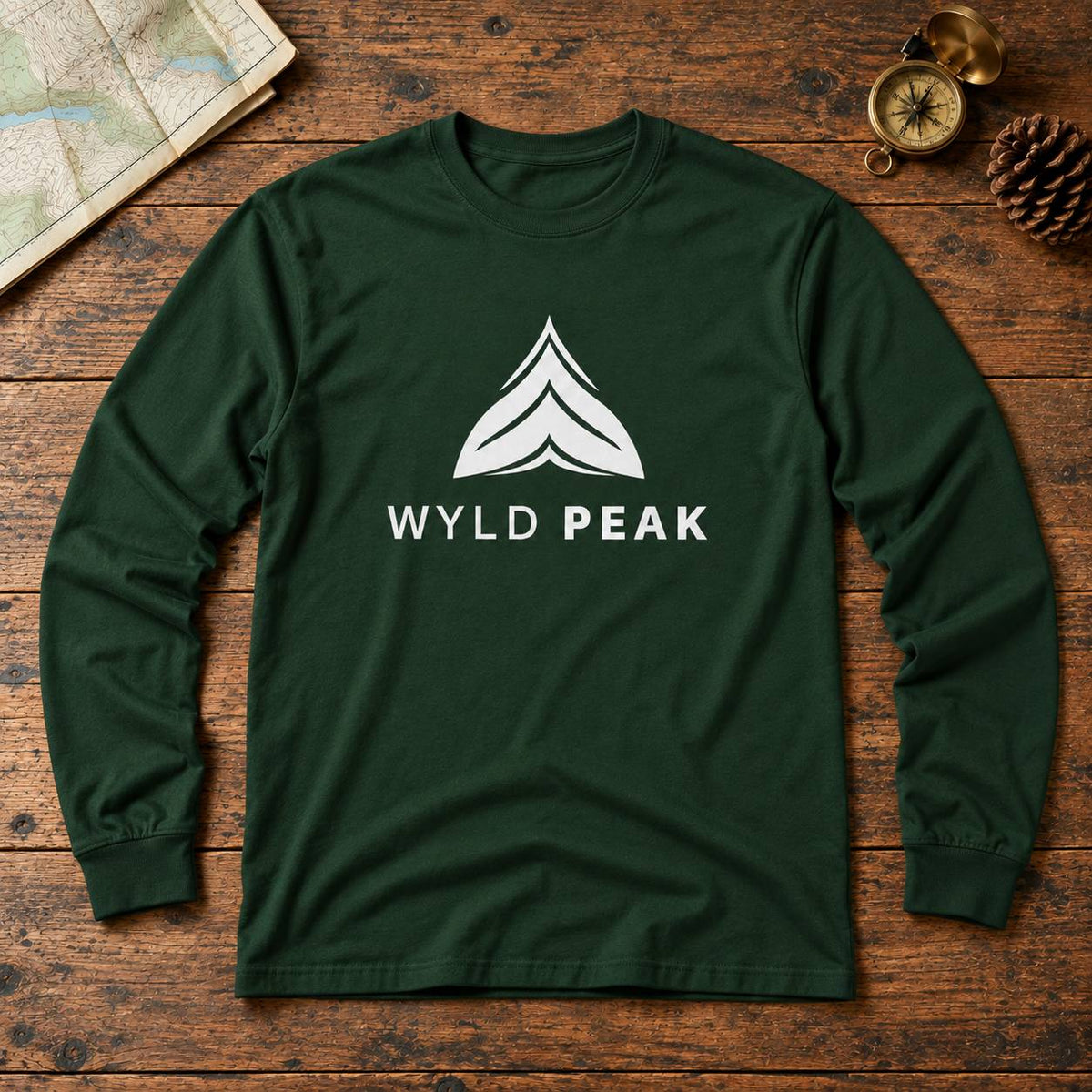 A dark green long-sleeve t-shirt with a white graphic logo and the text 'WYLD PEAK' printed on the chest, laid flat on a rustic wooden surface with a map, compass, and pinecone as props.