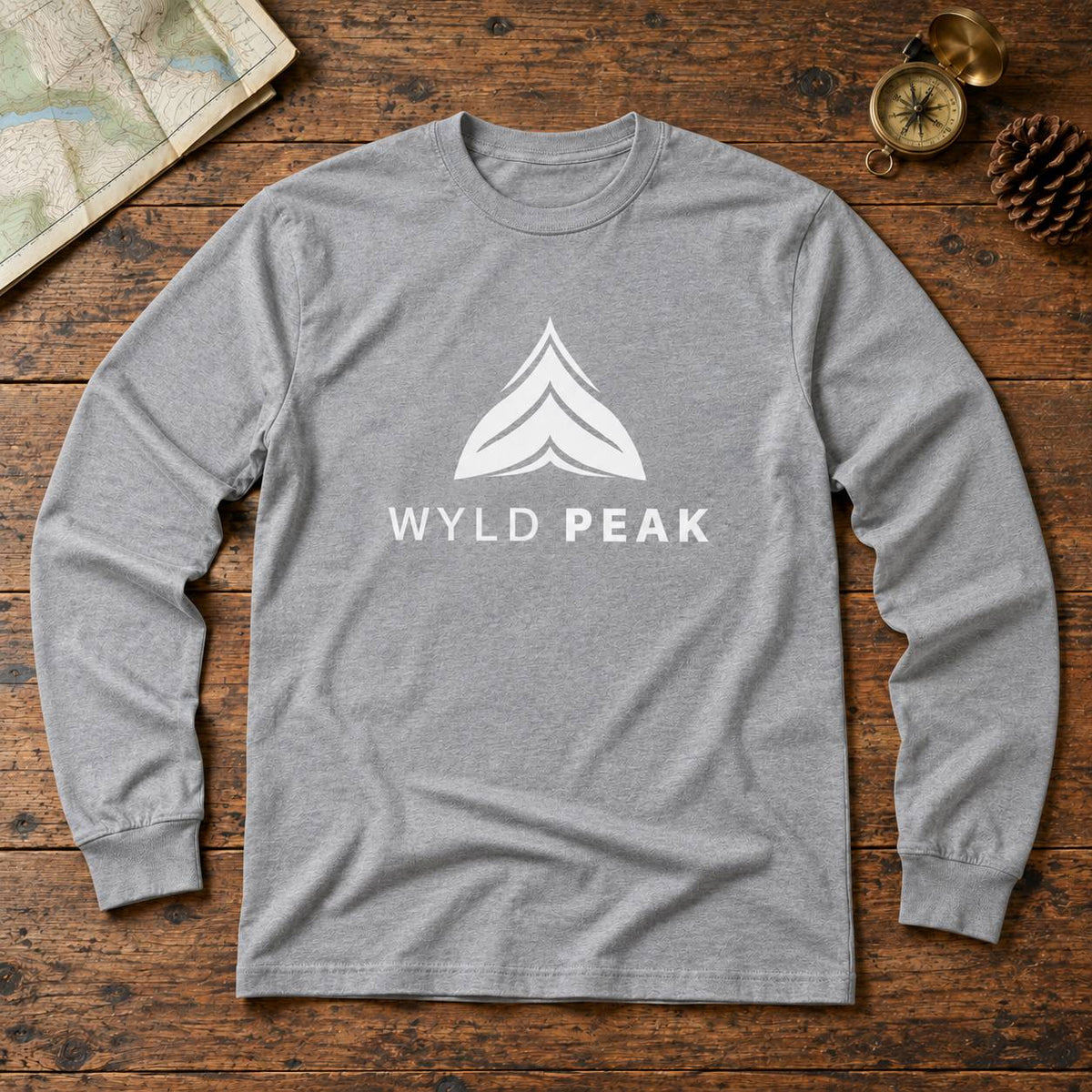 A heather gray long-sleeve t-shirt with a white graphic logo reading 'WYLD PEAK' and a mountain icon, laid flat on a rustic wooden surface with a map, compass, and pinecone as props.