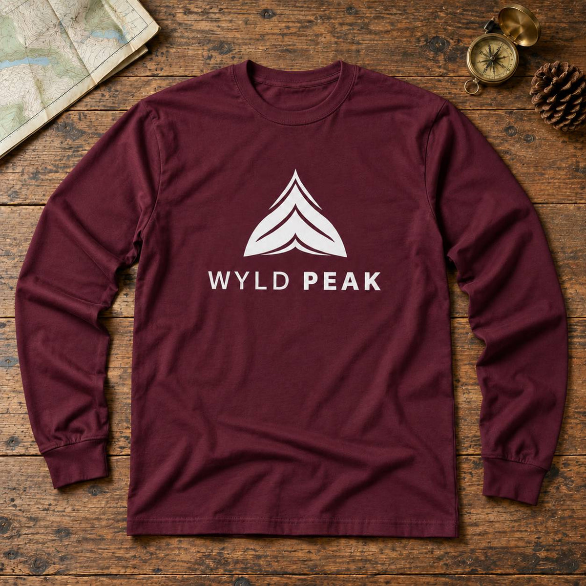 A maroon long-sleeve t-shirt with a white graphic logo and the text 'WYLD PEAK' on the chest, laid flat on a rustic wooden background with a map, compass, and pinecone as props.