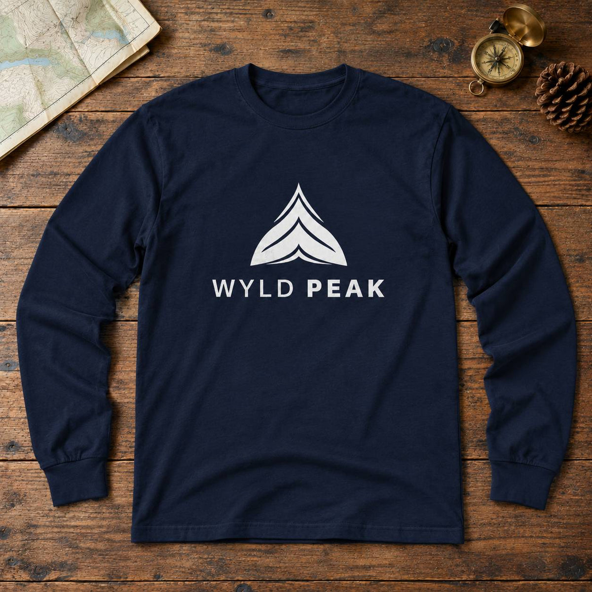 A navy blue long-sleeve t-shirt with a white graphic logo and the text 'WYLD PEAK' on the chest, laid flat on a rustic wooden background with a map, compass, and pinecone as props.