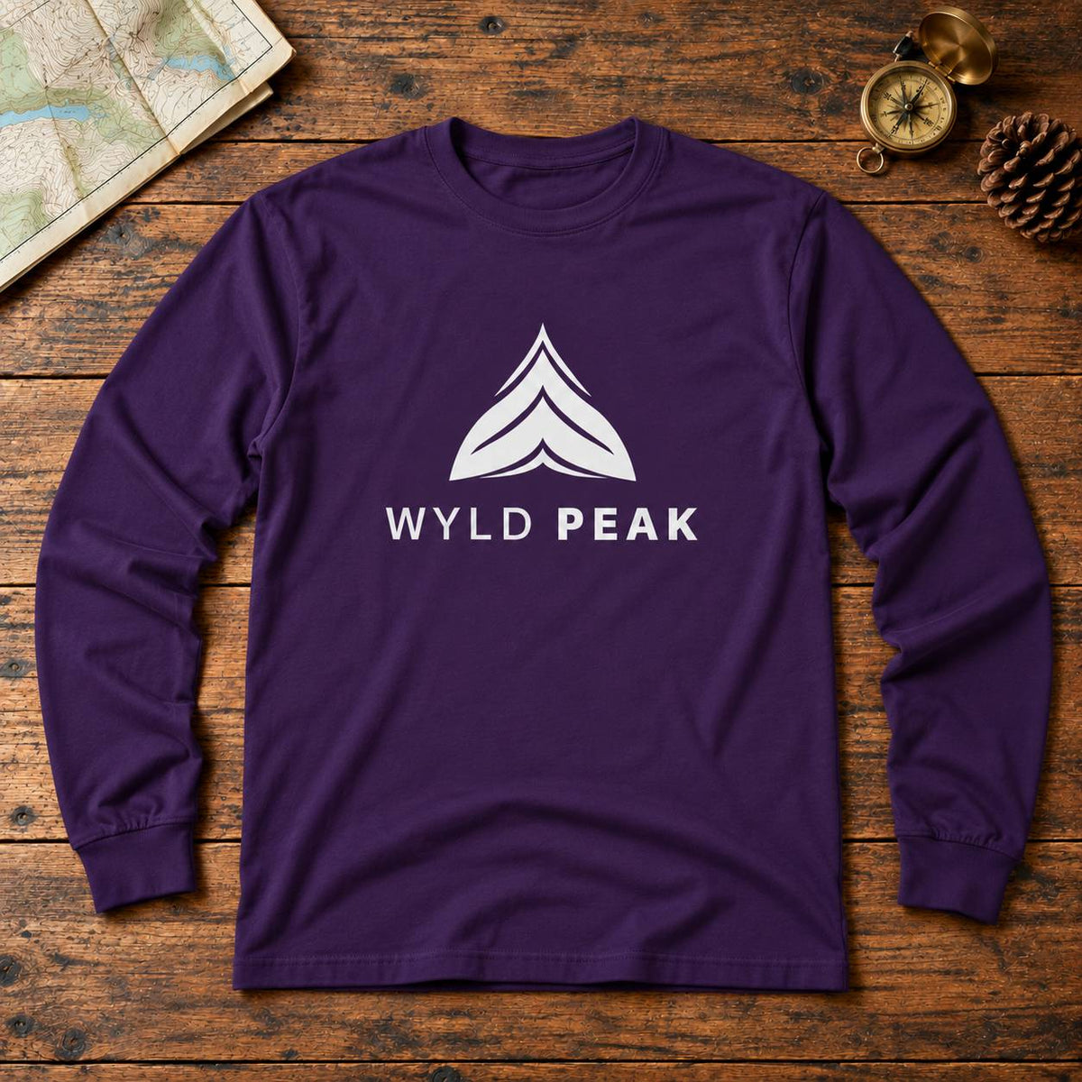 A purple long-sleeve t-shirt with a white 'WYLD PEAK' logo and mountain graphic on the chest, laid flat on a rustic wooden background with a map, compass, and pinecone as props.