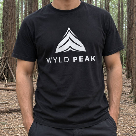 A man wearing a black crewneck t-shirt with a white graphic logo and the text 'WYLD PEAK' on the chest, standing in a forest of tall trees.