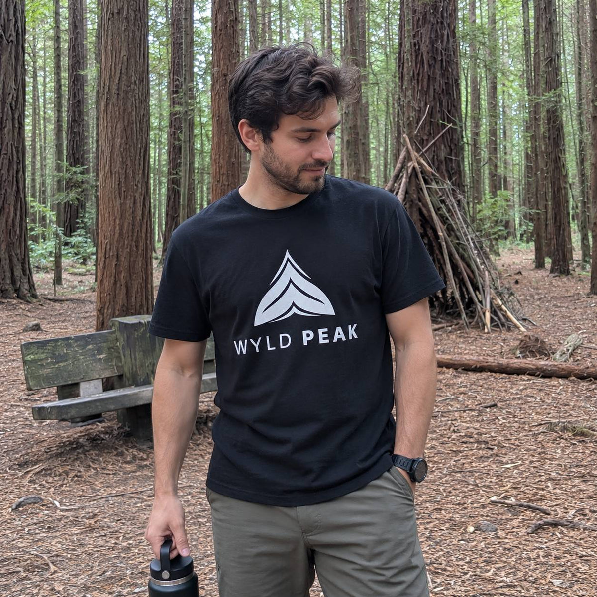 A man standing in a redwood forest wearing a black crewneck t-shirt with a white geometric mountain logo and the text 'WYLD PEAK' printed on the chest.