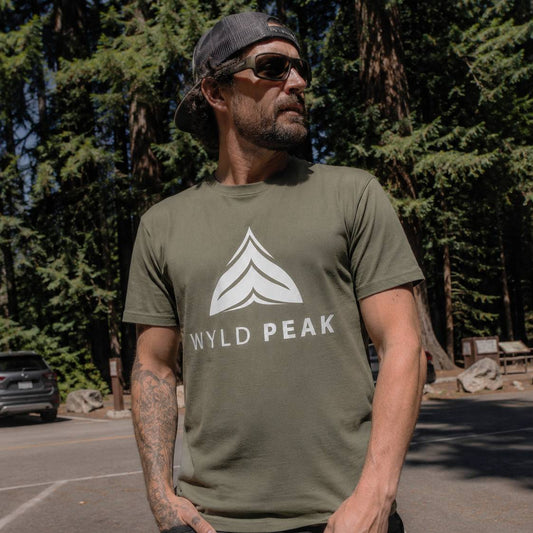 Man wearing a green 'Wyld Peak' t-shirt in a forest setting