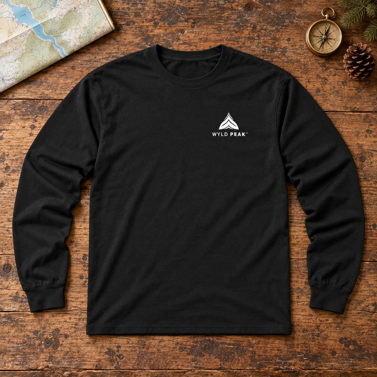 A black long-sleeve t-shirt with a white 'WYLD PEAK' logo on the left chest, laid flat on a rustic wooden surface with a map, compass, and pine cone as props.