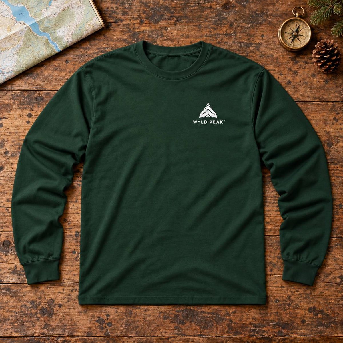 A dark forest green long-sleeve t-shirt with a small white 'WYLD PEAK' logo on the left chest, laid flat on a rustic wooden surface with outdoor-themed props like a map, compass, and pinecone.
