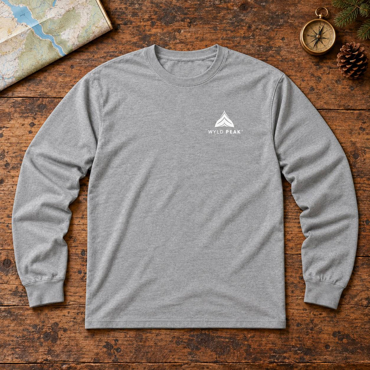 A heather gray long-sleeve t-shirt with a small white 'Wyld Peak' logo on the left chest, laid flat on a rustic wooden surface with a map, compass, and pinecone as props.