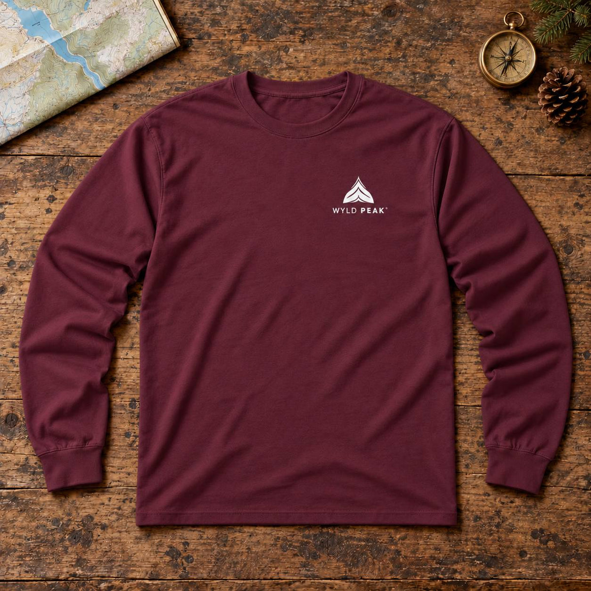 A burgundy long-sleeve t-shirt with a white 'WYLD PEAK' logo on the left chest, laid flat on a rustic wooden surface with outdoor-themed props like a map, compass, and pinecone.