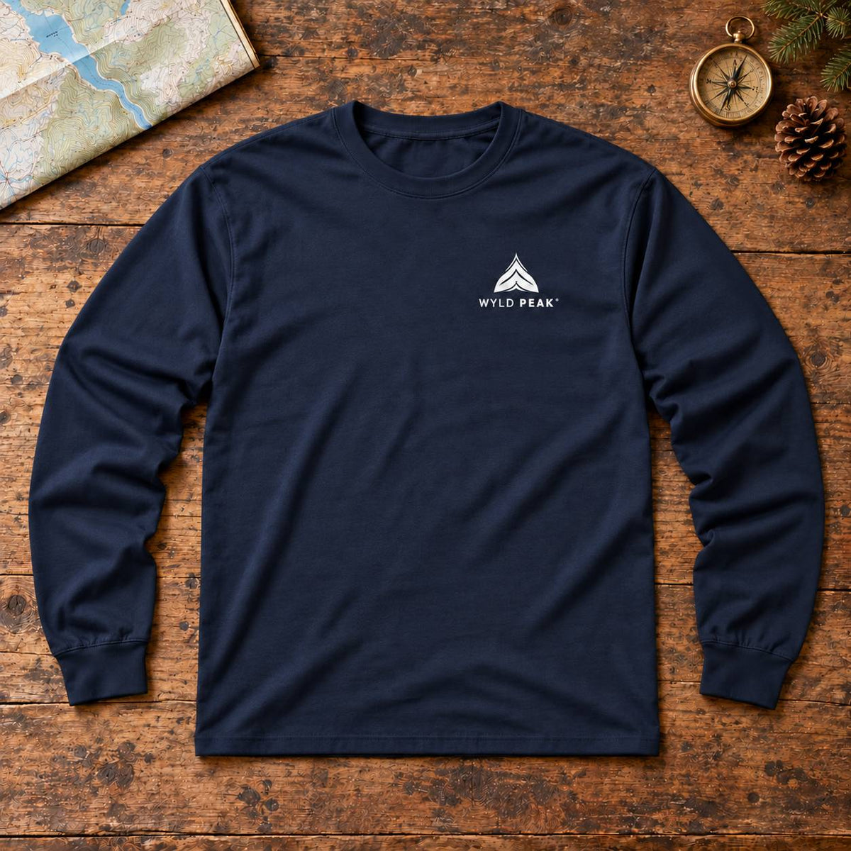 A navy blue long-sleeve t-shirt with a white 'WYLD PEAK' logo on the left chest, laid flat on a rustic wooden surface with a map, compass, and pinecone as props.