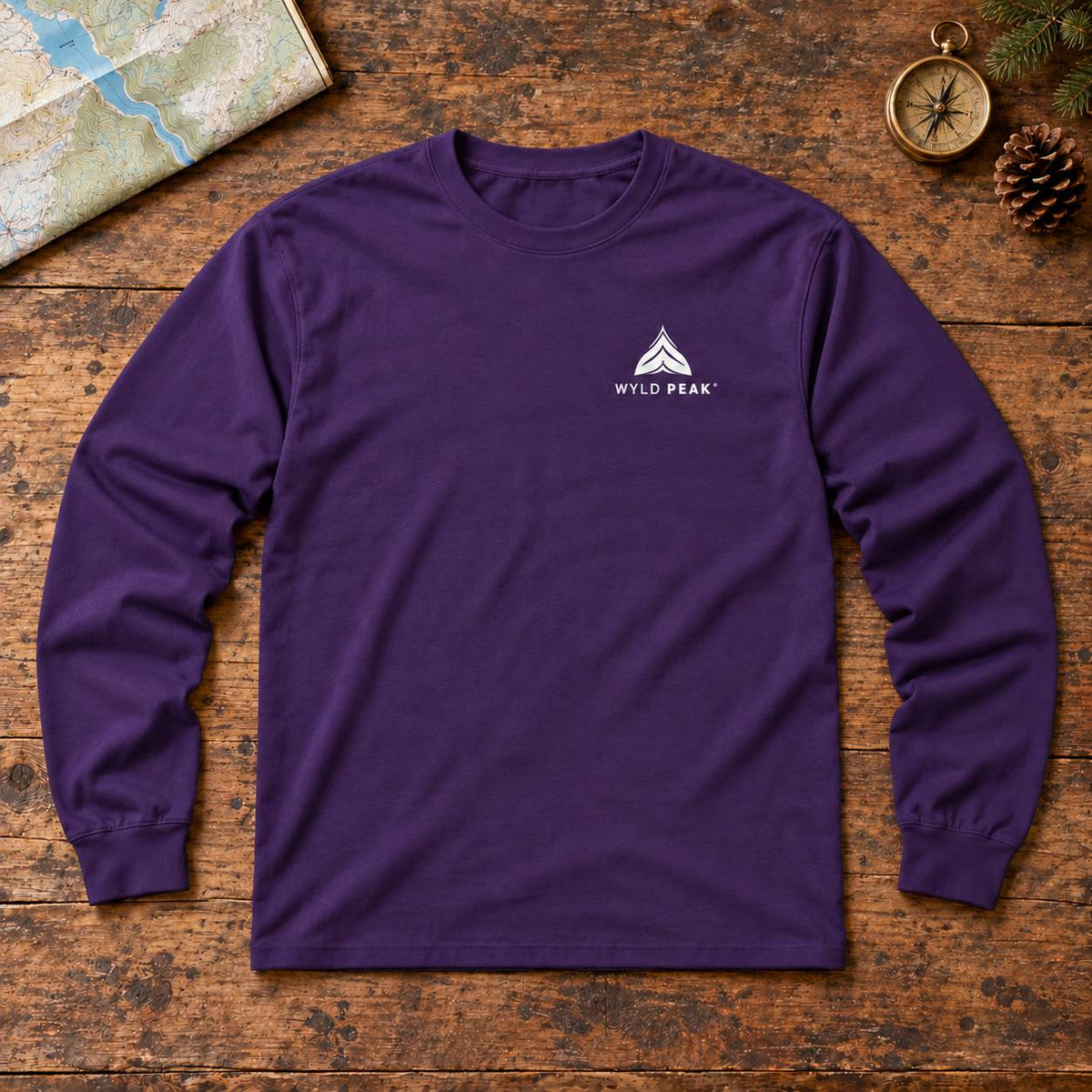 A purple long-sleeve t-shirt with a white 'WYLD PEAK' logo on the left chest, laid flat on a rustic wooden surface with a map, compass, and pine cone as props.