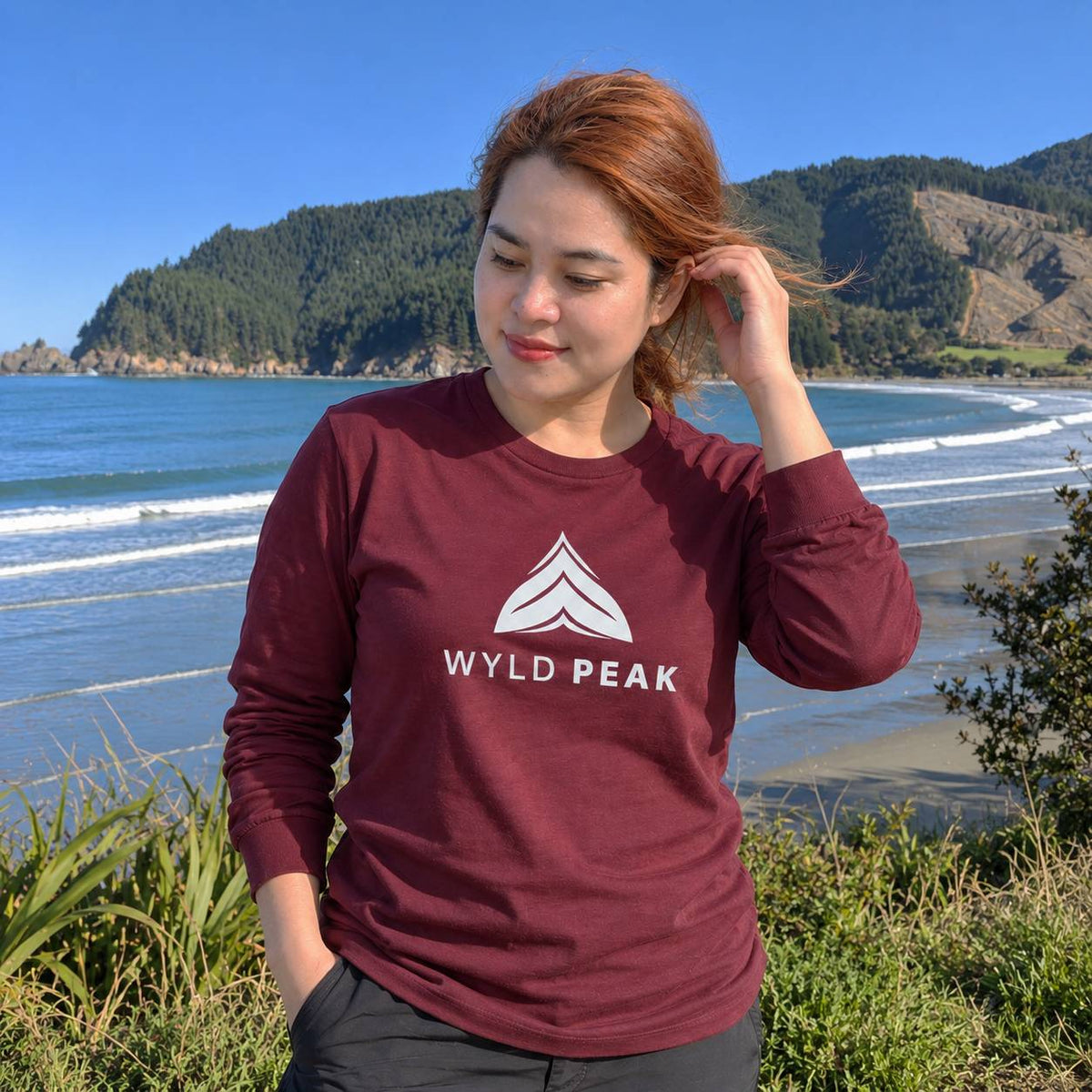 A woman wearing a maroon long-sleeve t-shirt with a white 'WYLD PEAK' logo stands on a grassy hill overlooking a scenic ocean and mountain landscape.