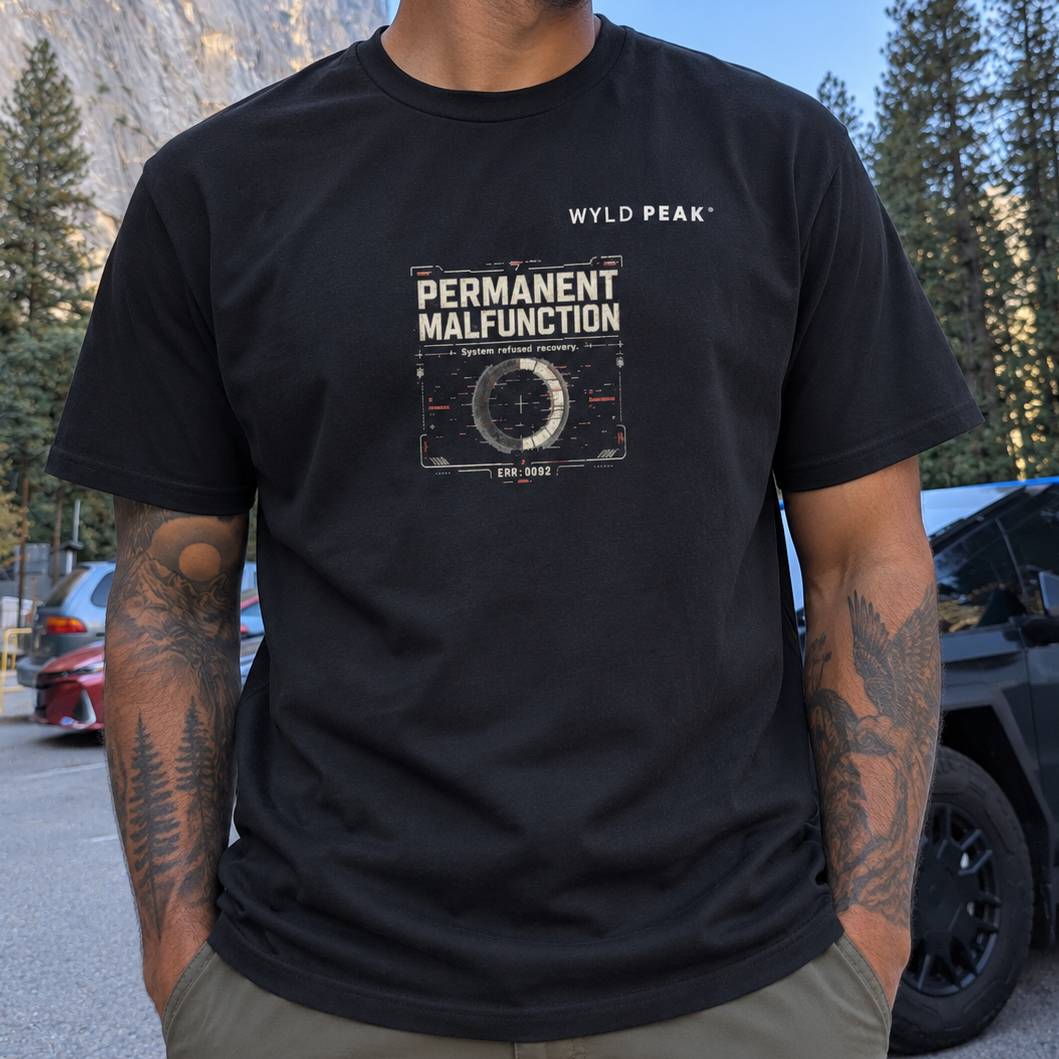 A man wearing a black graphic t-shirt with a distressed white print that reads 'WYLD PEAK' and 'PERMANENT MALFUNCTION'. The background shows an outdoor mountain setting with trees and a vehicle.