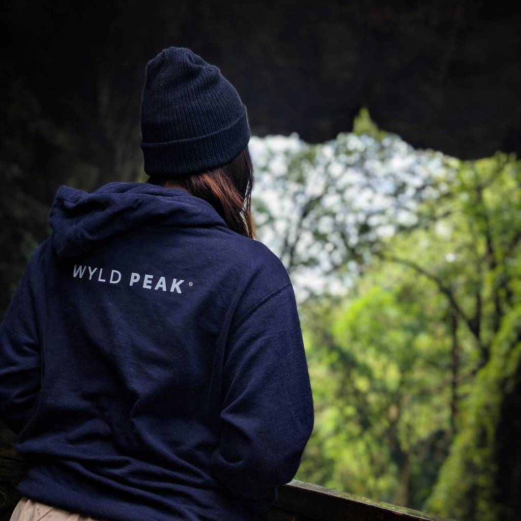 Person wearing a navy hoodie with 'Wyld Peak' logo, looking at trees