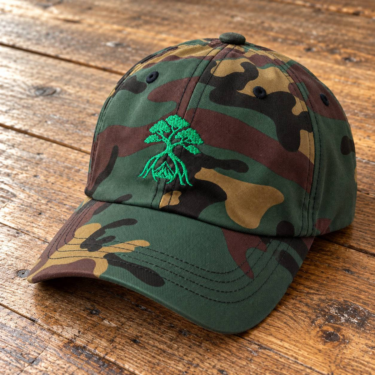 A woodland camouflage baseball cap with a green embroidered tree graphic on the front, resting on a rustic wooden surface.