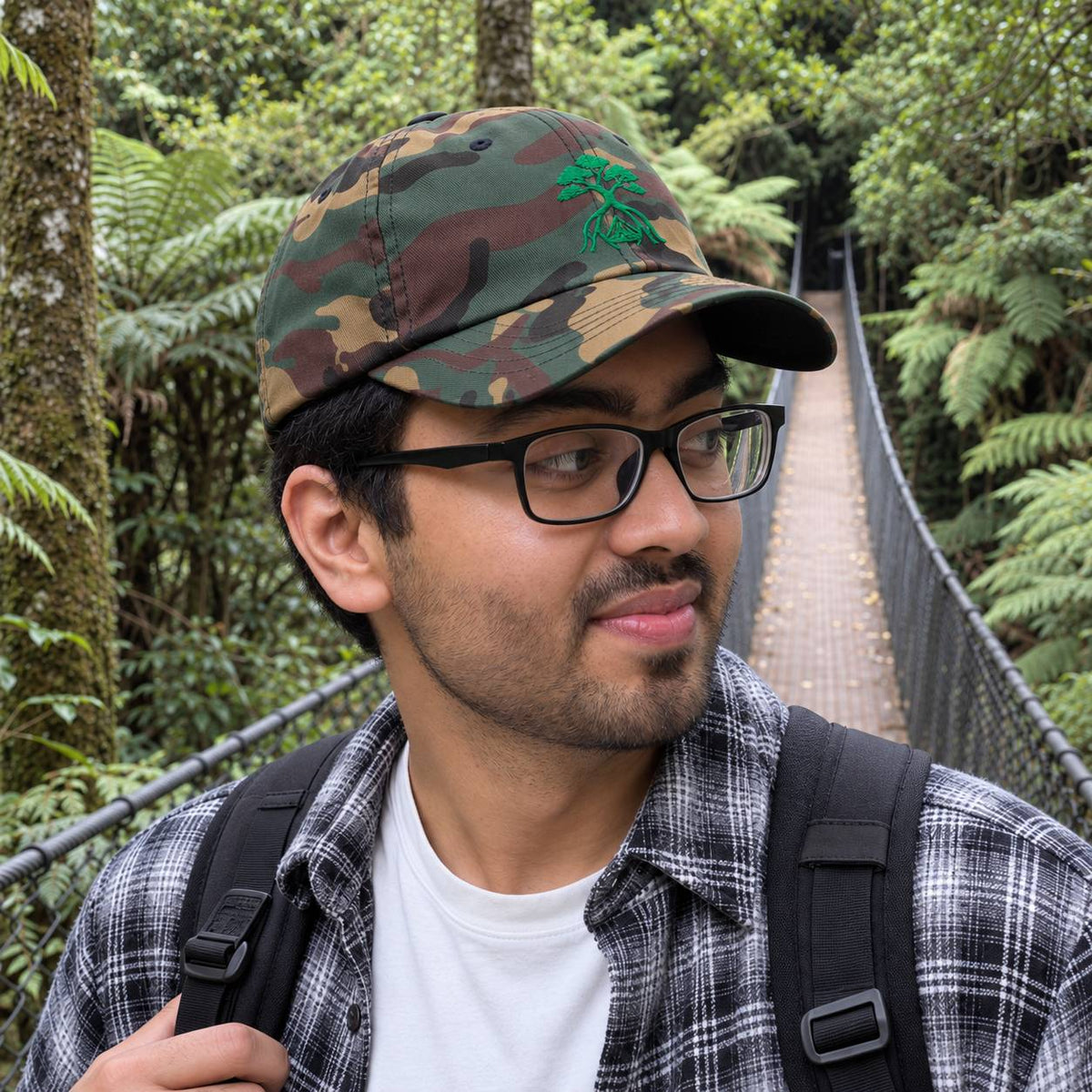 A man wearing a green camouflage baseball cap with a green tree logo, black-rimmed glasses, a plaid flannel shirt, and a backpack, standing on a suspension bridge surrounded by lush green forest.