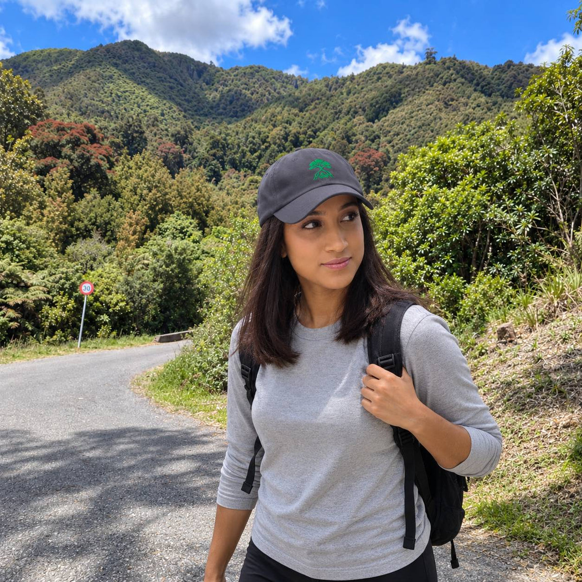 A woman hiking on a path wearing a gray long-sleeve shirt, a dark gray baseball cap with a green logo, and a black backpack, with a lush green mountain landscape in the background.
