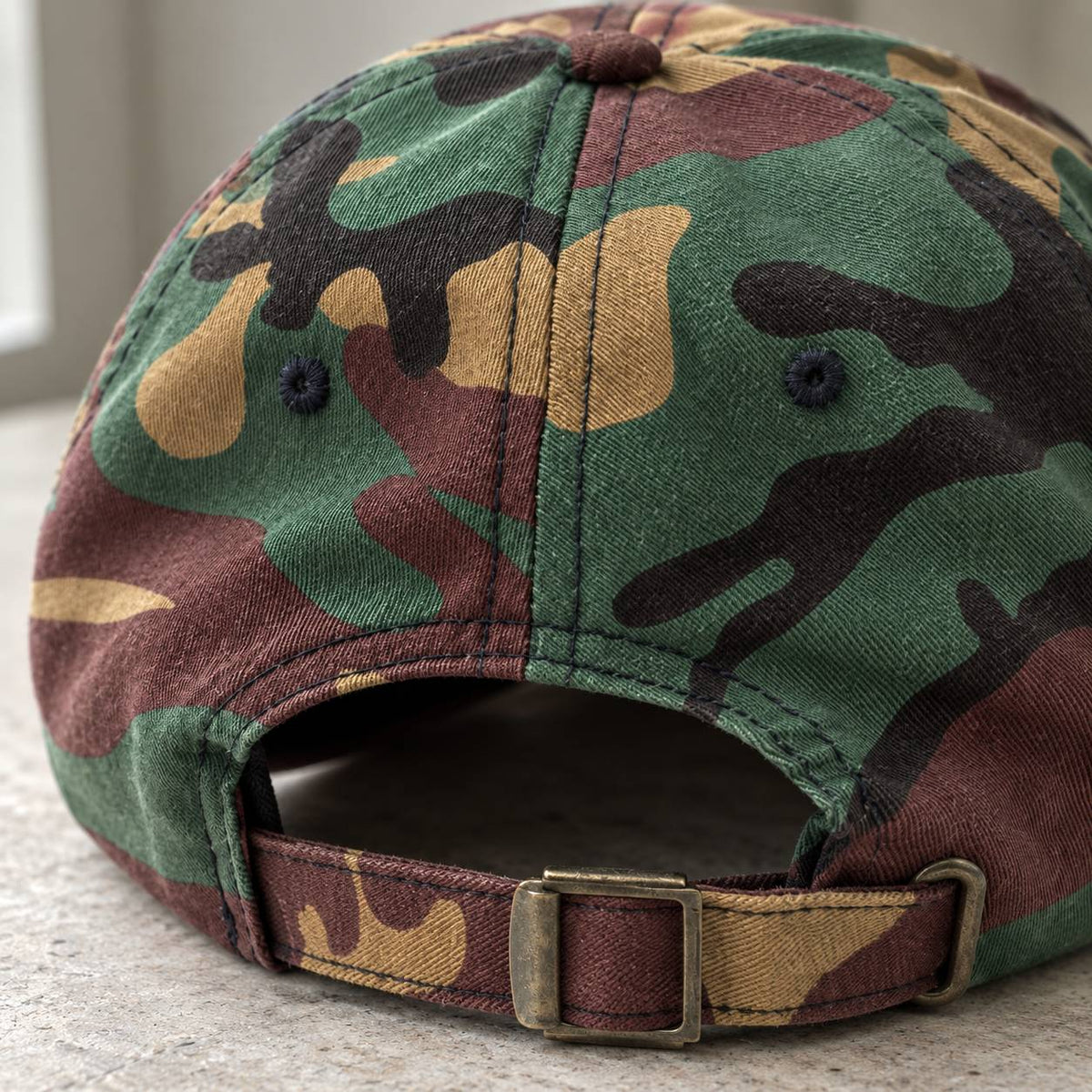 Close-up of the back of a camouflage baseball cap with an adjustable fabric strap and metal buckle, shown on a light stone surface.