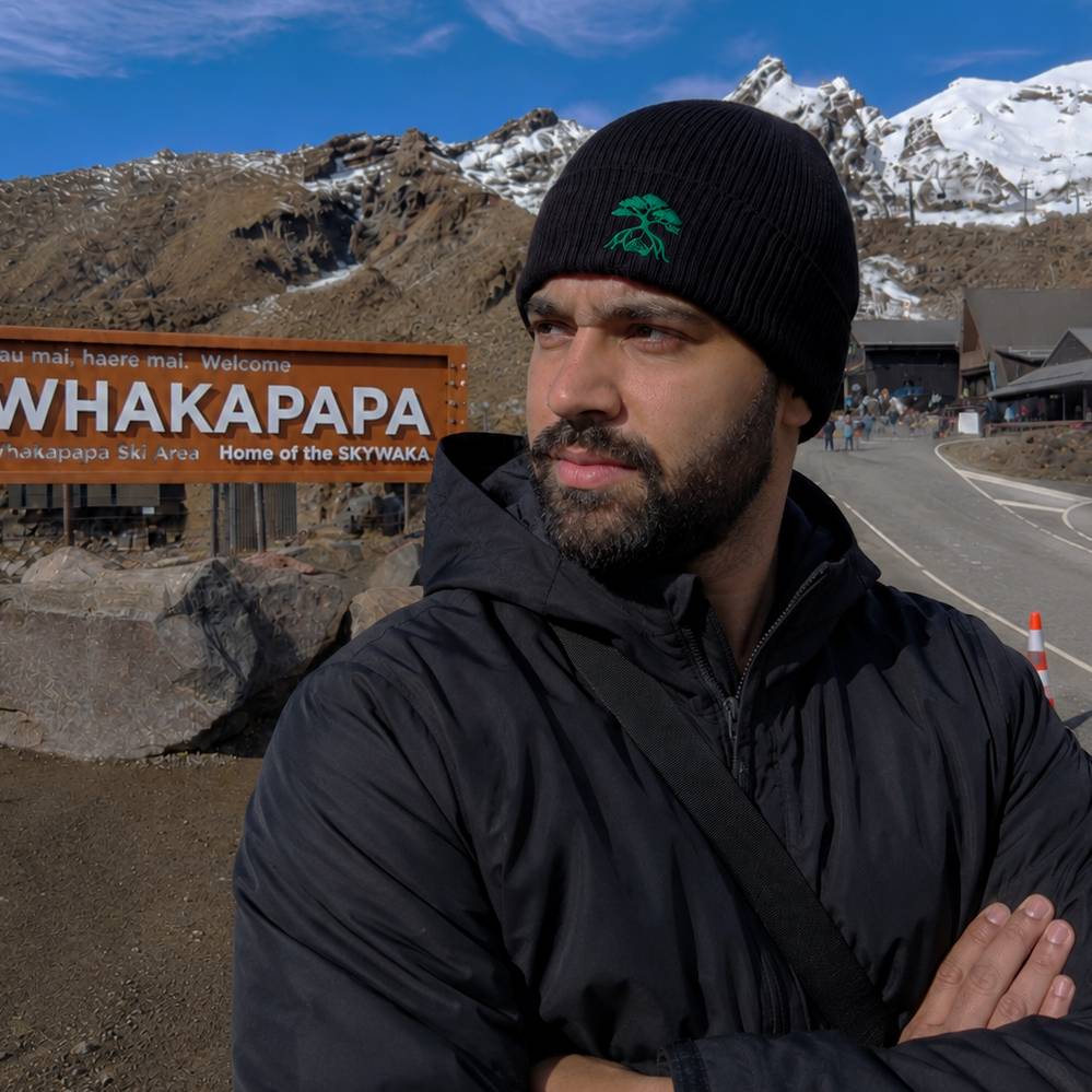 A man wearing a black knit beanie with a green tree logo and a black jacket stands in front of a Whakapapa sign and snowy mountains under a blue sky.