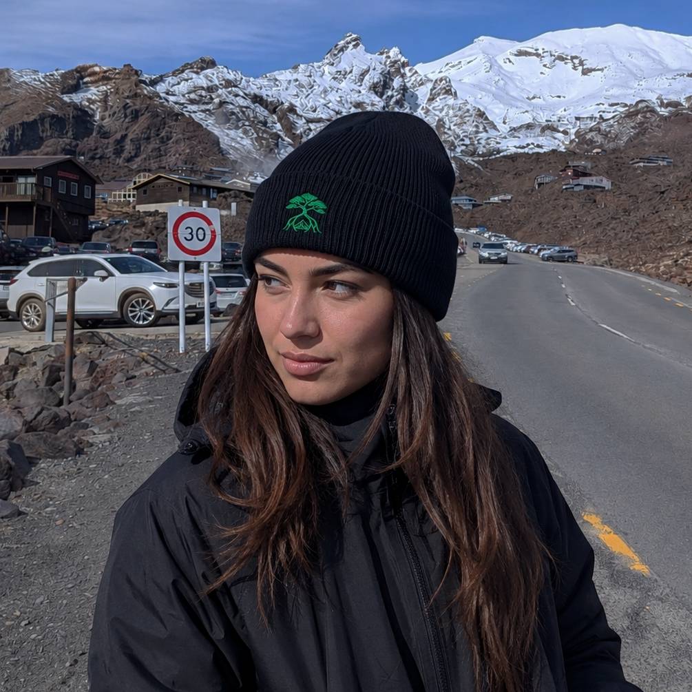 A woman wearing a black ribbed knit beanie with a green tree logo and a black jacket stands on a mountain road with snow-capped peaks in the background.