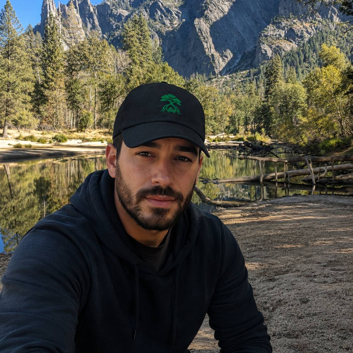 A man wearing a black baseball cap with a green mangrove tree logo and a black hoodie, taking a selfie in front of a mountain and lake landscape.