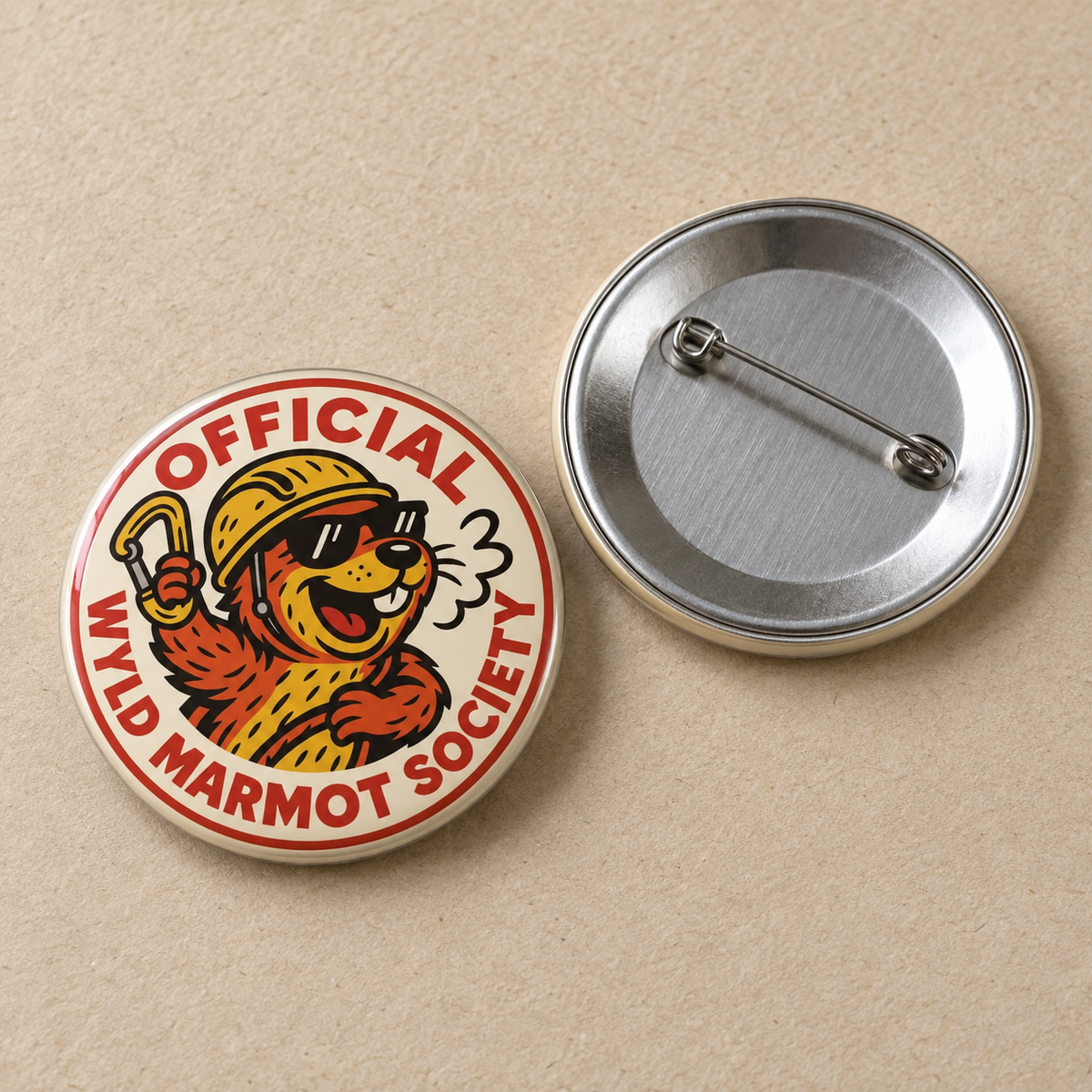 Two round pin-back buttons on a beige textured background. The left button shows the front design with a cartoon marmot wearing a yellow hard hat and sunglasses, surrounded by the text 'OFFICIAL WYLD MARMOT SOCIETY' in red.