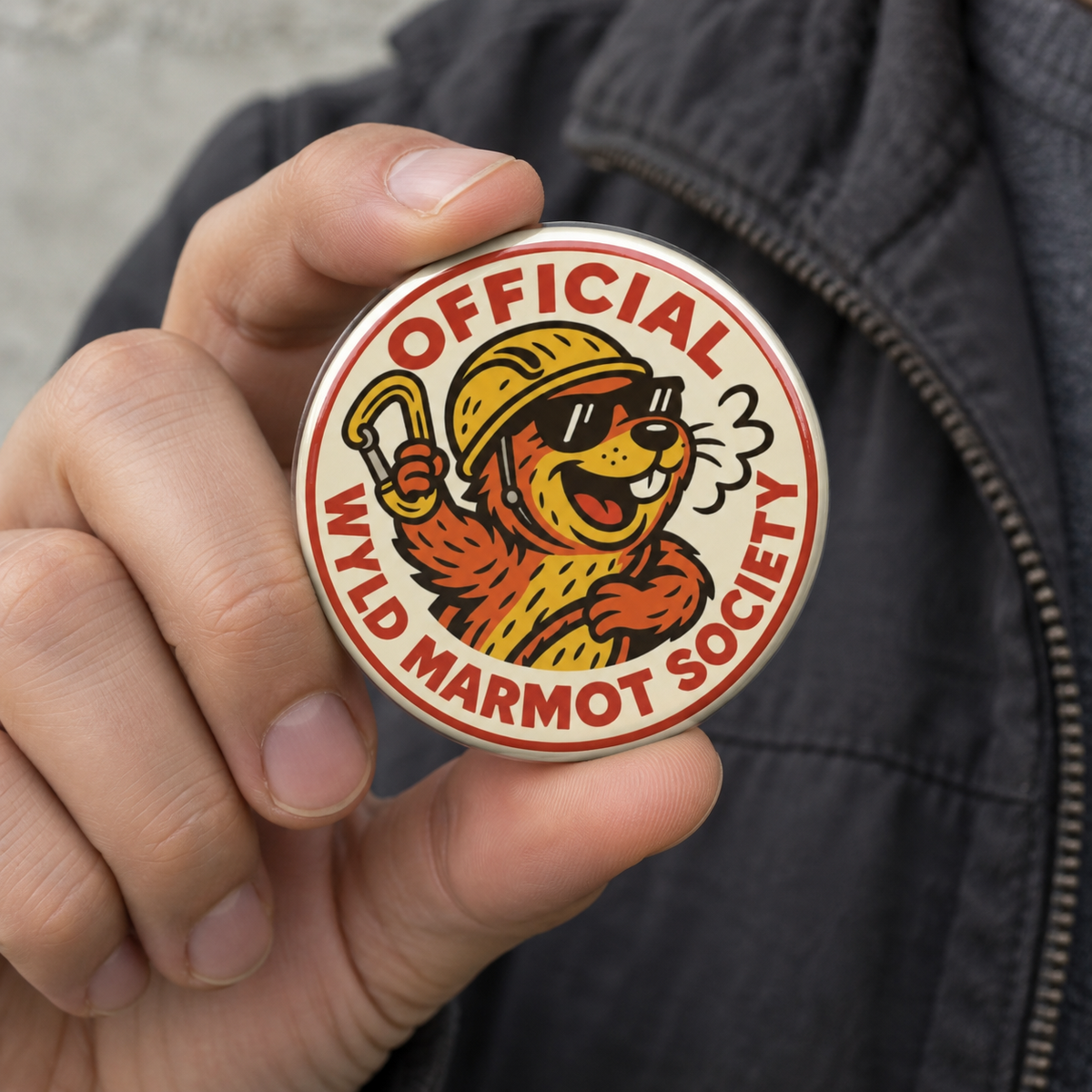 A close-up of a hand holding a round enamel pin that reads 'OFFICIAL WYLD MARMOT SOCIETY' with a cartoon marmot wearing a yellow helmet and sunglasses, against a dark gray jacket background.