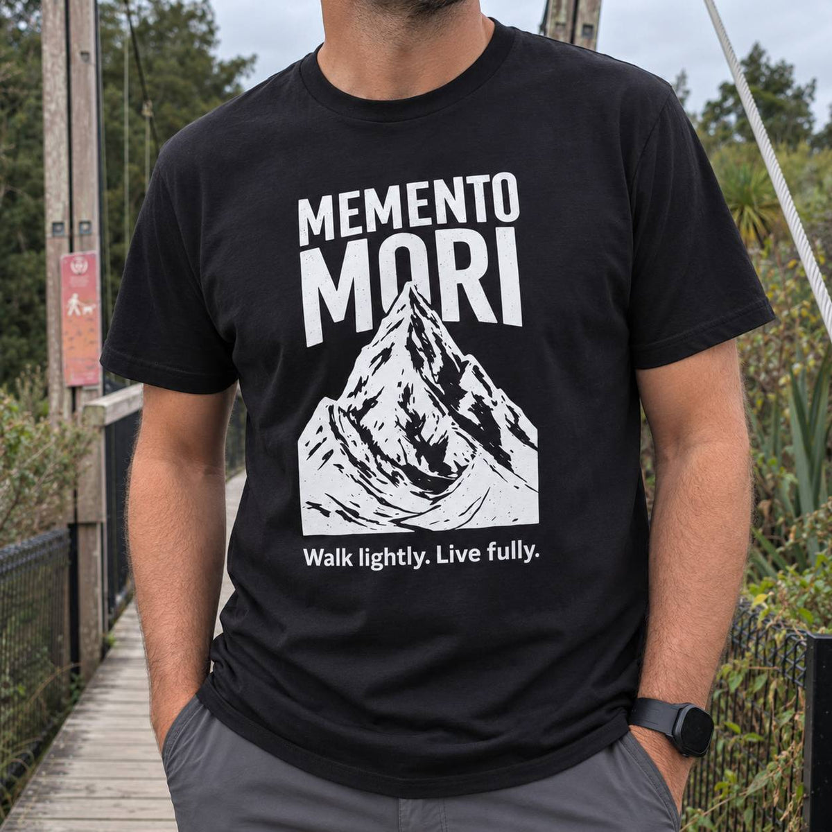 A man wearing a black t-shirt with a white graphic print that reads 'MEMENTO MORI' above a mountain illustration and the phrase 'Walk lightly. Live fully.
