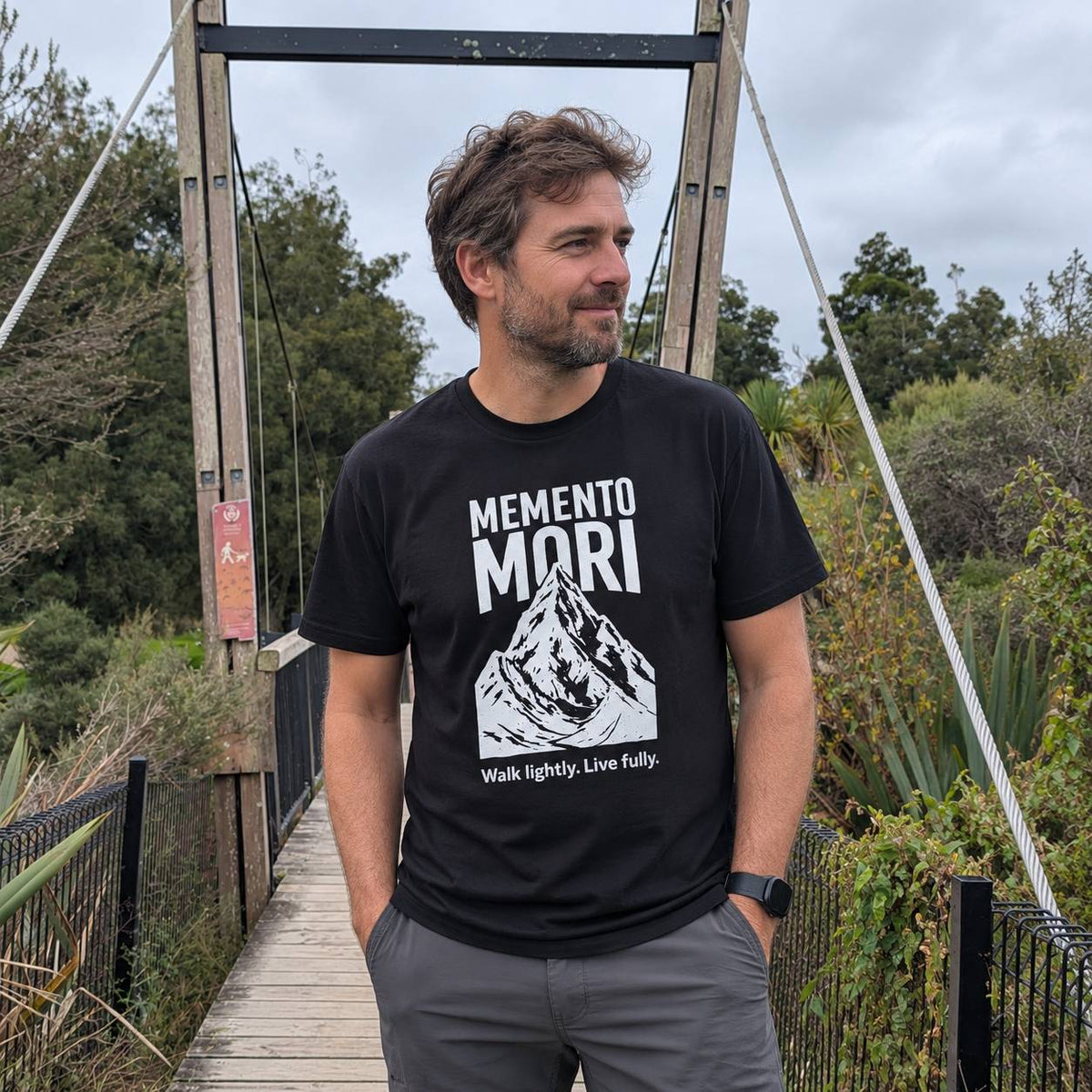 A man standing on a wooden suspension bridge wearing a black t-shirt with a white graphic print that reads 'MEMENTO MORI' and 'Walk lightly. Live fully.