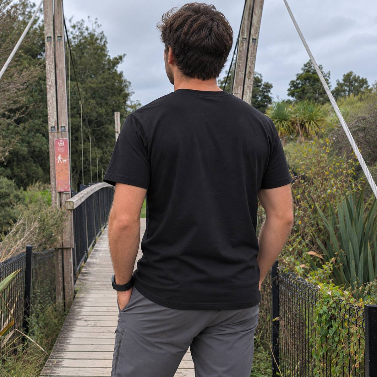 A man viewed from behind wearing a black short-sleeve crewneck t-shirt and gray pants, standing on a wooden suspension bridge surrounded by green foliage under an overcast sky.