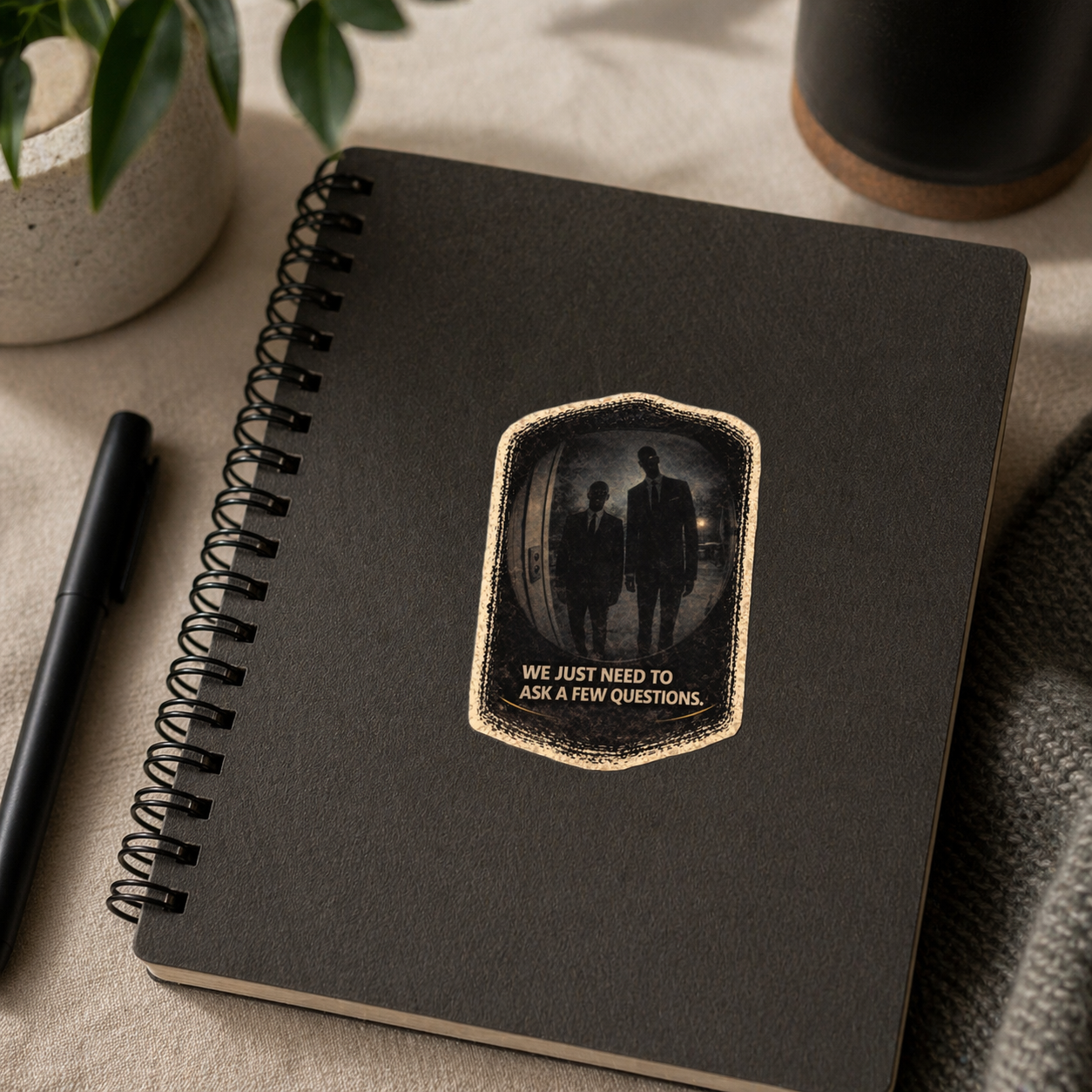Black notebook with a decorative patch featuring two people and text, placed on a textured surface with a pen and plant.