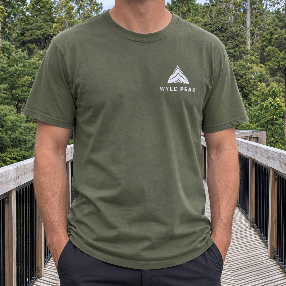 A man wearing an olive green crewneck t-shirt with a white 'WYLD PEAK' logo on the left chest, standing on a wooden boardwalk with a forest background.