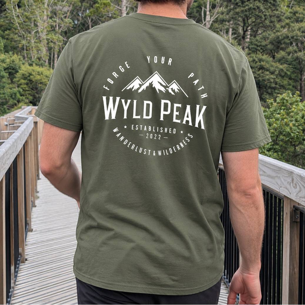 A man viewed from behind wearing an olive green t-shirt with a white graphic print on the back that reads 'WYLD PEAK' with mountain silhouettes and outdoor text. He is standing on a wooden boardwalk with trees in the background.