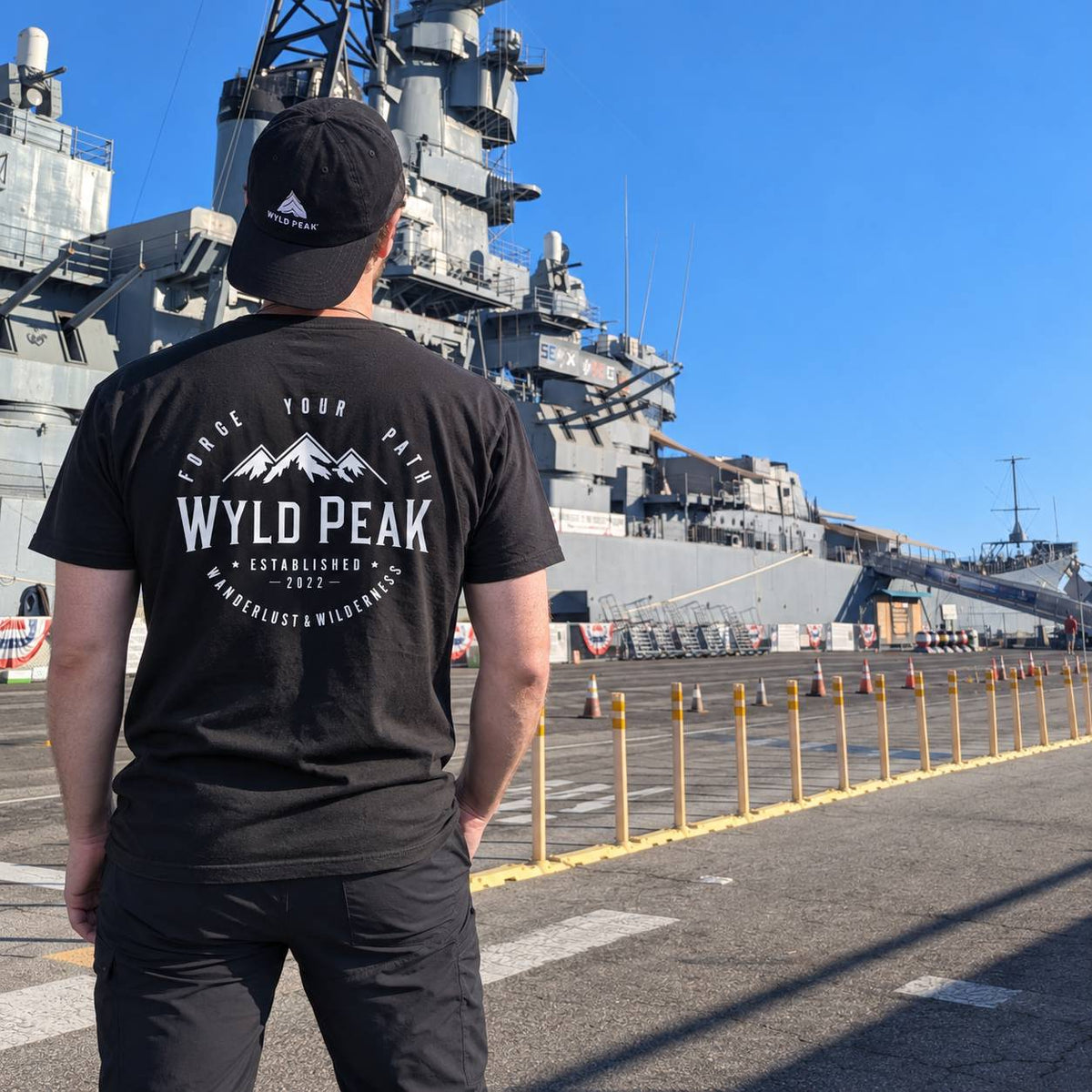 A man viewed from behind wearing a black t-shirt and black cap, standing on a dock with a large gray naval ship in the background under a clear blue sky. The t-shirt features a white graphic with mountains and the text 'WYLD PEAK'.