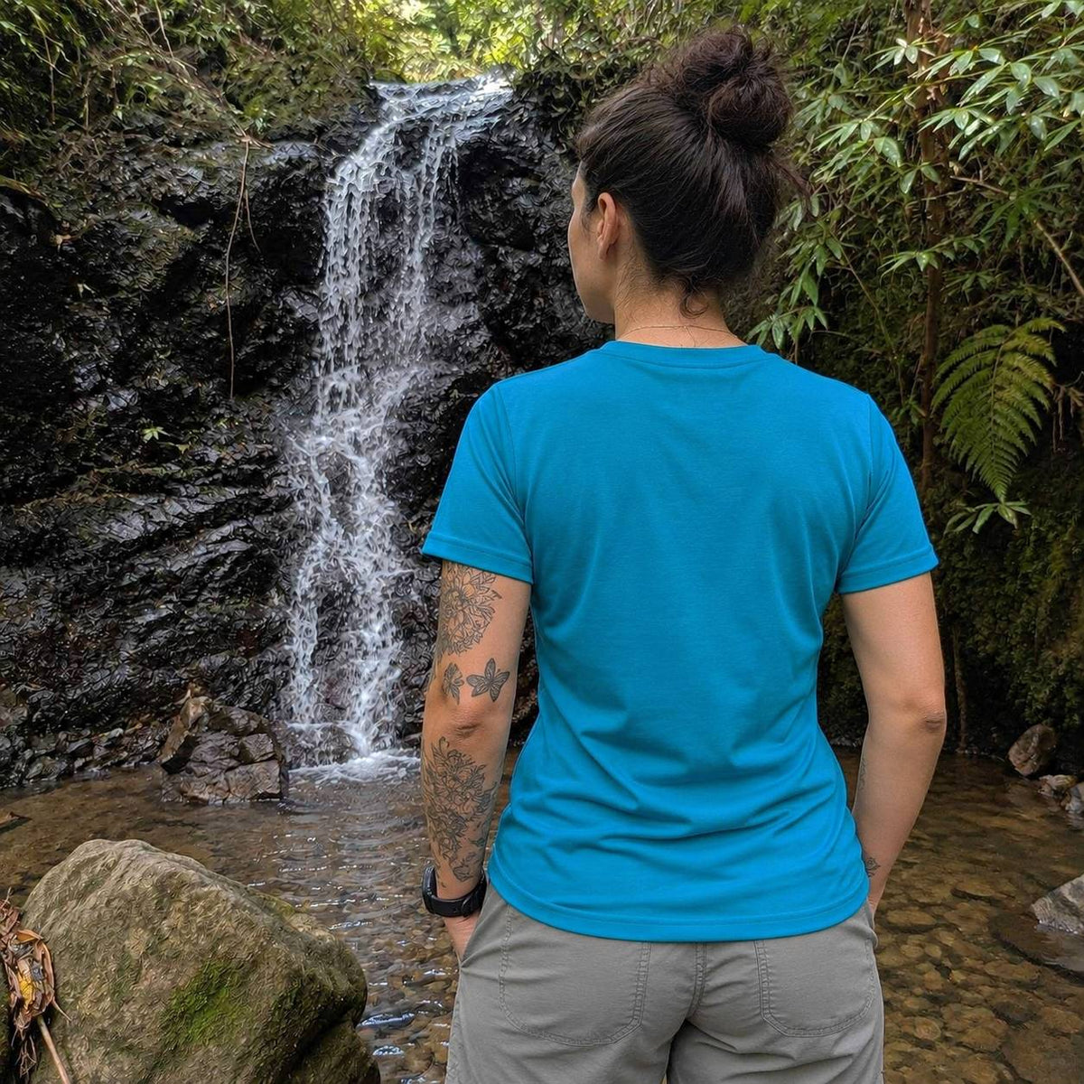 A woman viewed from behind wearing a bright blue crewneck t-shirt and khaki pants, standing in front of a small waterfall in a lush green outdoor setting.
