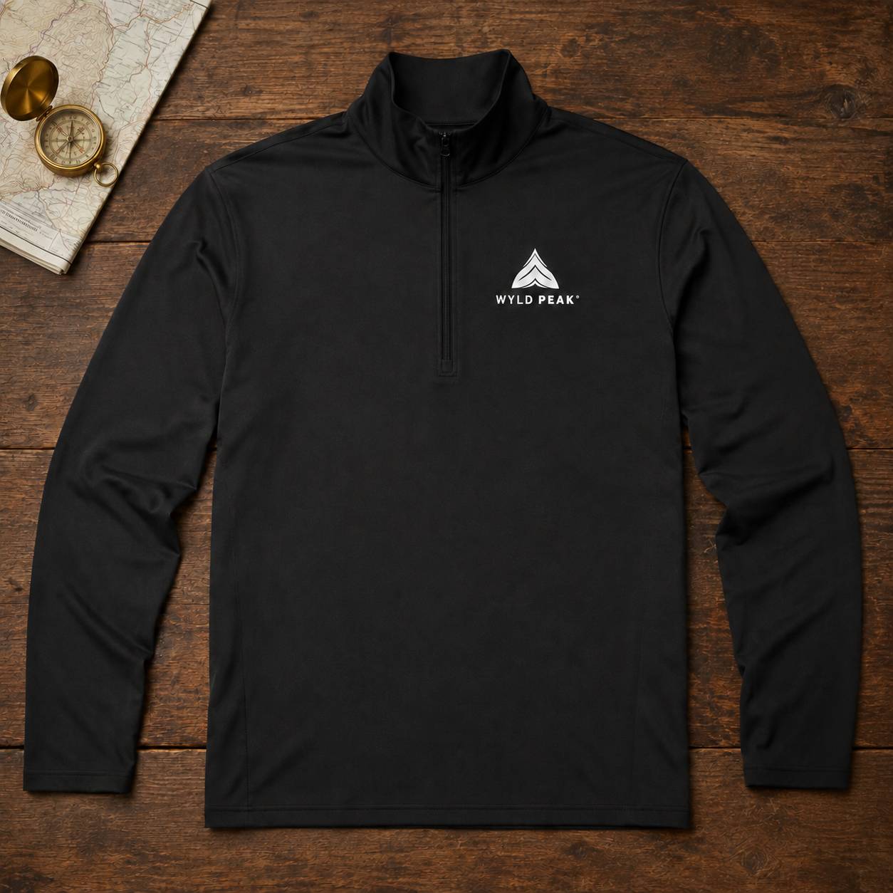 A black long-sleeve quarter-zip pullover laid flat on a rustic wooden surface, with a small white Wild Peak logo printed on the left chest. A vintage map and compass are partially visible in the top-left corner as styled props.