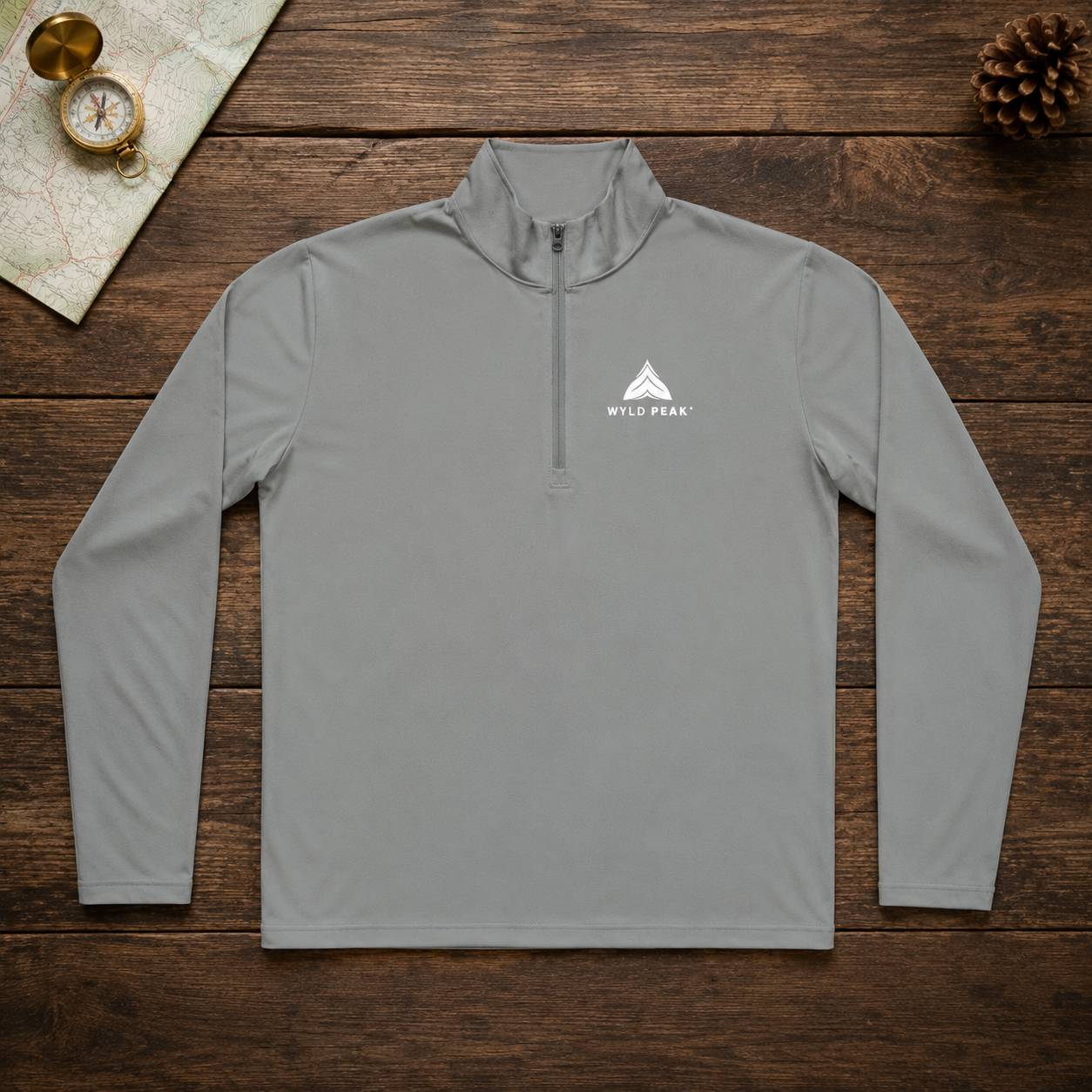 A light gray long-sleeve quarter-zip pullover with a white 'Wild Peak' mountain logo on the left chest, laid flat on a dark wood background with a map, brass compass, and pinecone as props.