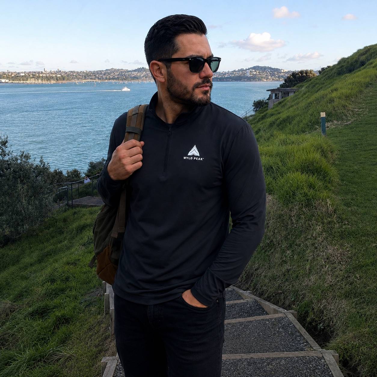 A man wearing a black long-sleeve quarter-zip pullover with a white 'WYLD PEAK' logo on the chest, standing outdoors on stone steps with a scenic ocean and grassy hill in the background.
