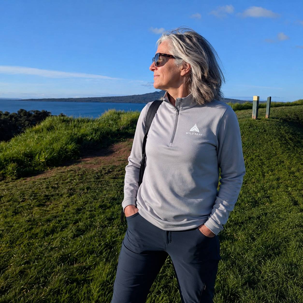 A woman with gray hair and sunglasses stands outdoors on a grassy hill overlooking water, wearing a light gray quarter-zip pullover with a white mountain logo and the text 'WYLD PEAK' on the chest, along with dark pants.