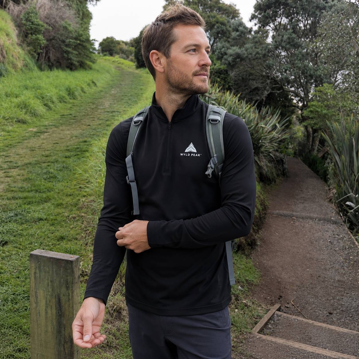 A man hiking on a grassy path wearing a black long-sleeve quarter-zip athletic top with a white Anta Peak logo on the chest, along with a gray backpack.