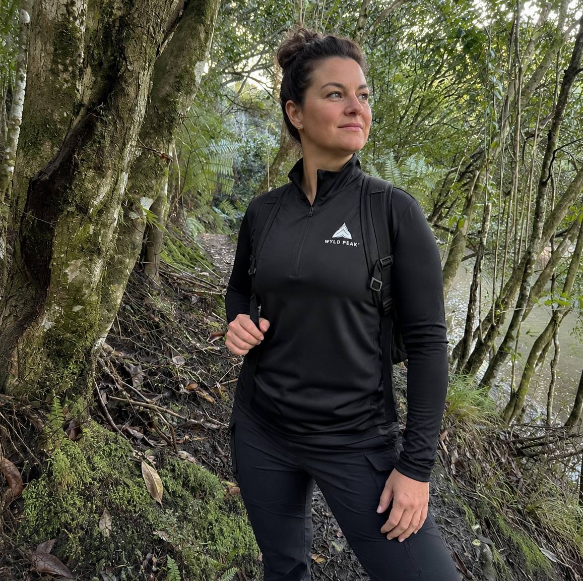 A woman wearing a black long-sleeve athletic quarter-zip top with a white 'WYLD PEAK' logo on the chest, standing in a mossy forest environment.