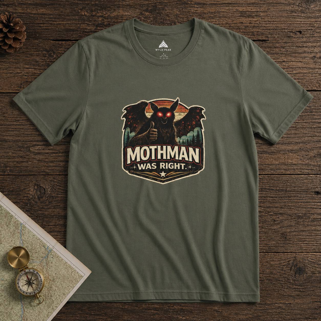 Green t-shirt with Mothman graphic and 'Mothman Was Right' text on a wooden surface.